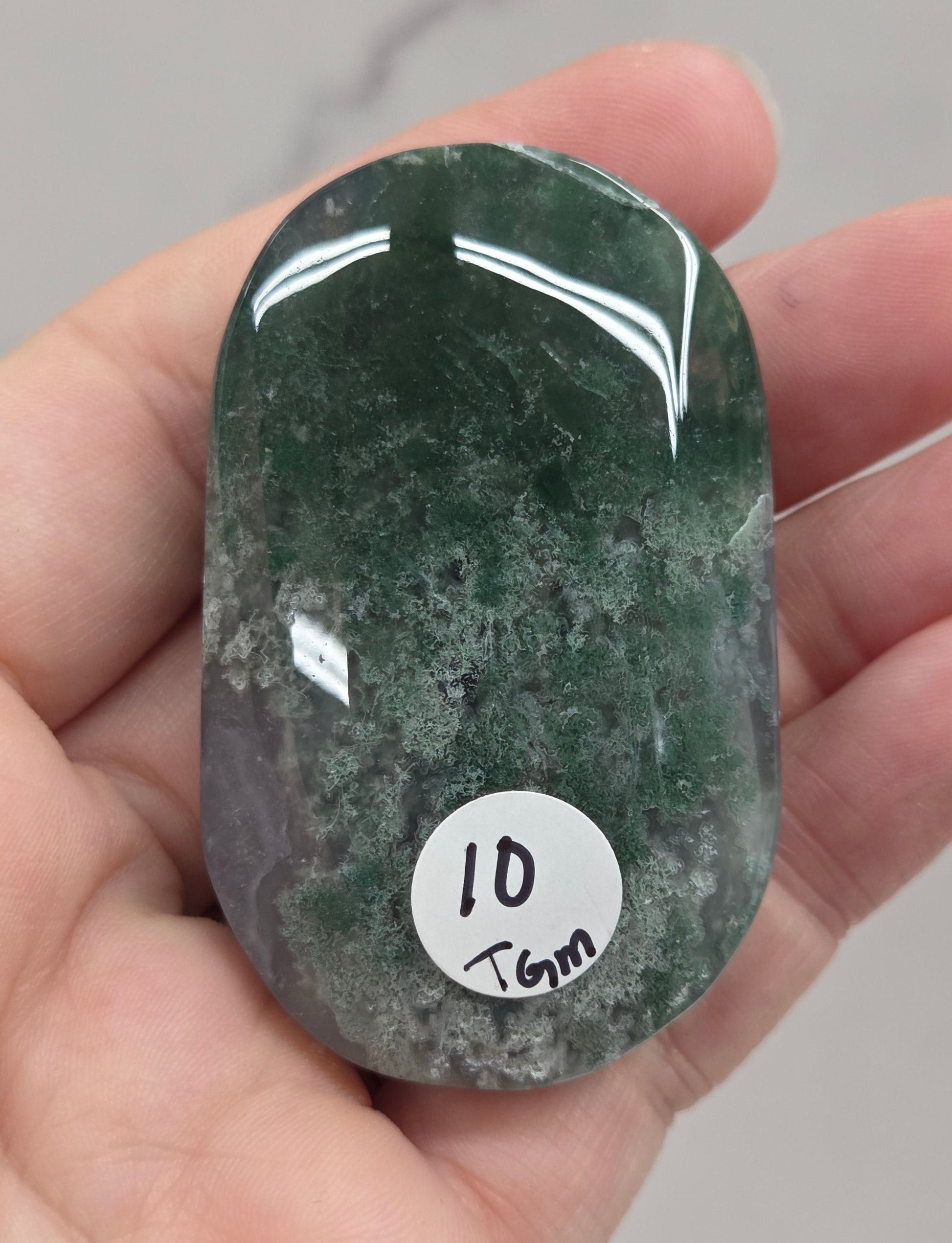 A hand holding a polished dark green stone pendant with a small white circular sticker that reads '10 mm'.