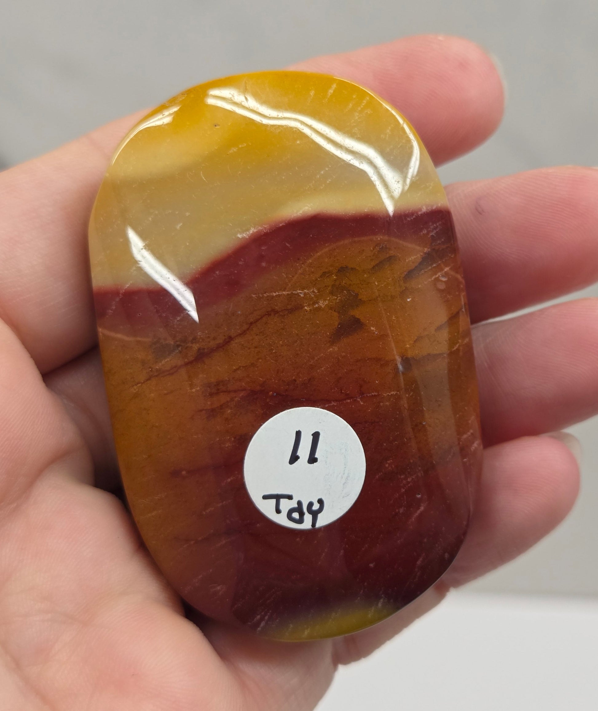 A hand holding a polished, rounded stone with a marbled pattern of yellow, amber, and reddish-brown. A small white sticker with handwritten text is attached to the stone.