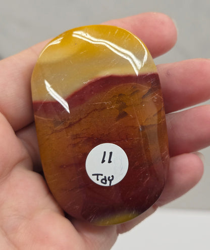 A hand holding a polished, rounded stone with a marbled pattern of yellow, amber, and reddish-brown. A small white sticker with handwritten text is attached to the stone.
