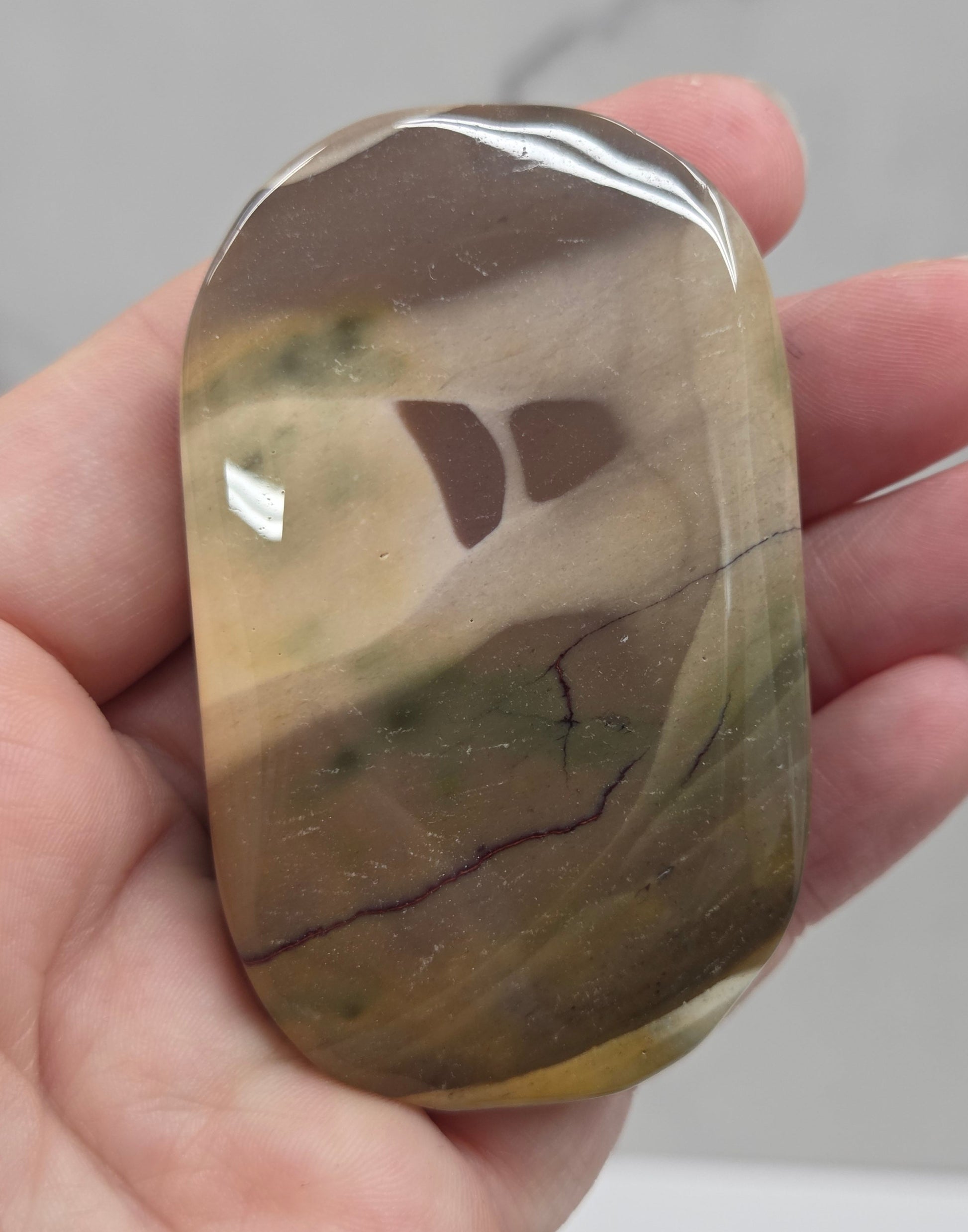 A polished stone with a rounded, elongated shape held in a person's hand. The stone has a marbled pattern of brown, beige, and olive green with a glossy finish.