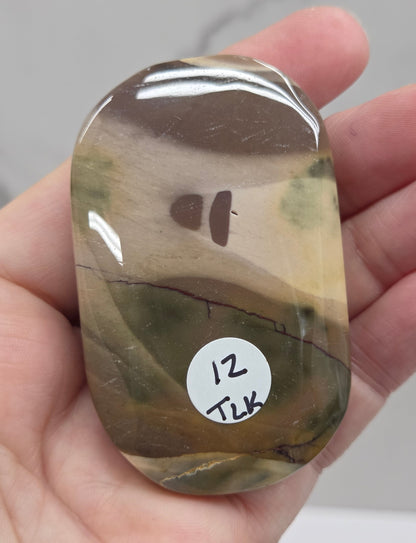 A hand holding a polished, rounded stone with a marbled pattern of brown, olive green, and beige, featuring a small white sticker with handwritten text on the right side.