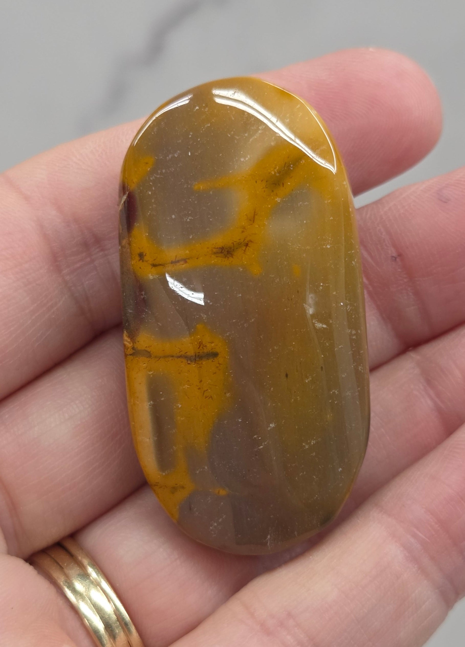 A polished brown and yellow stone held in a person's hand, with a gold ring on one finger against a light marble-like background.