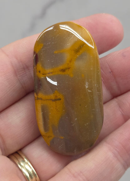 A polished brown and yellow stone held in a person's hand, with a gold ring on one finger against a light marble-like background.