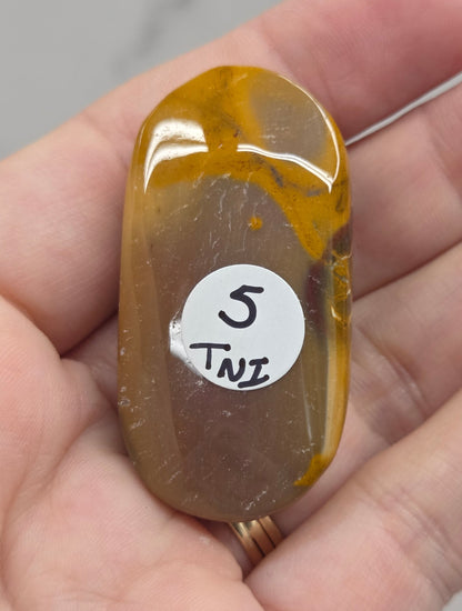 A polished oval-shaped brown and amber stone pendant or cabochon held in a person's hand, with a small white sticker labeled '5 TWE' on the front.