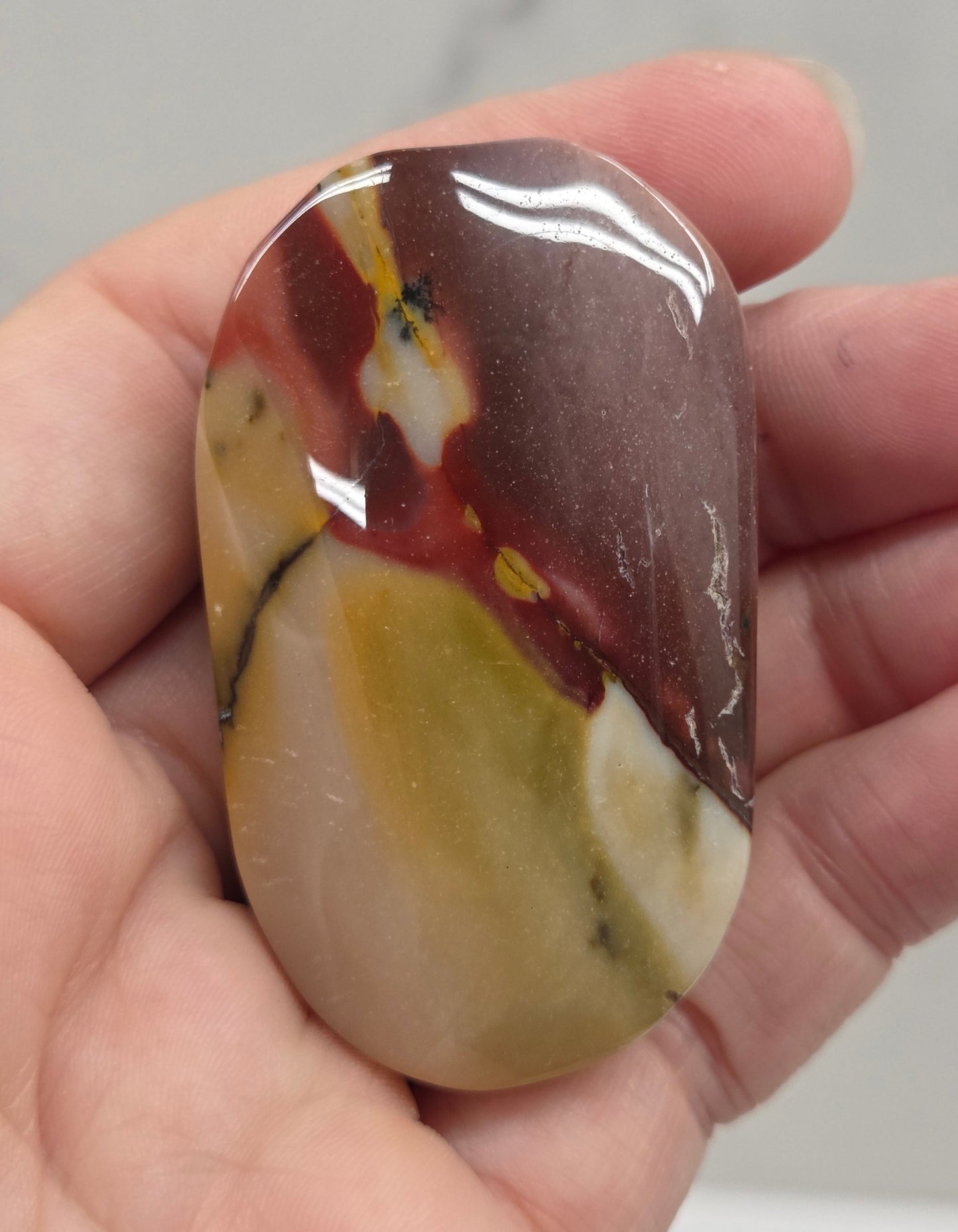 A polished, smooth stone with a marbled pattern of brown, cream, yellow, and red, held in a person's hand against a light gray marble background.