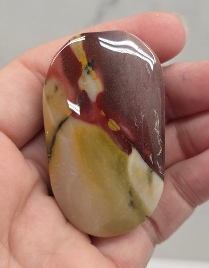 A polished, smooth stone with a marbled pattern of brown, cream, yellow, and red, held in a person's hand against a light gray marble background.