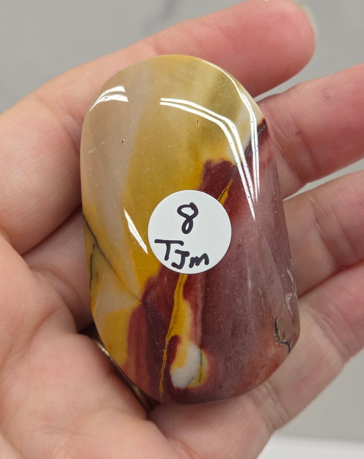 A polished tumbled stone with marbled yellow, brown, and cream patterns, held in a person's hand. A small white circular sticker with handwritten text is attached to the stone.