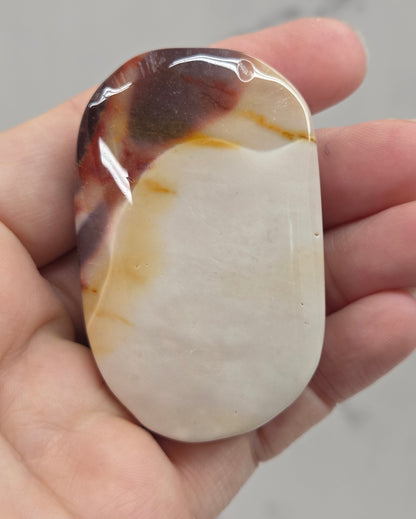 A hand holding a polished, smooth stone pendant with a creamy off-white center and dark brown to black marbled edges.