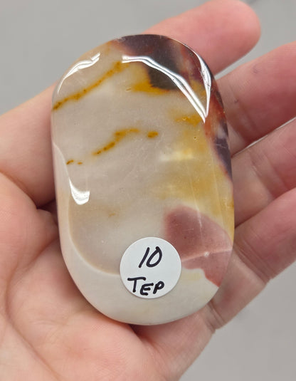 A hand holding a polished, tumbled stone with natural marbled patterns in cream, yellow, and dark brown. A small white sticker on the stone reads '10 Tep'.