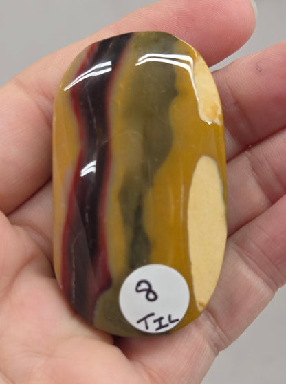 A hand holding a polished, rounded stone with horizontal bands of yellow, olive green, black, and reddish-brown, featuring a small white sticker with handwritten text on the right side.