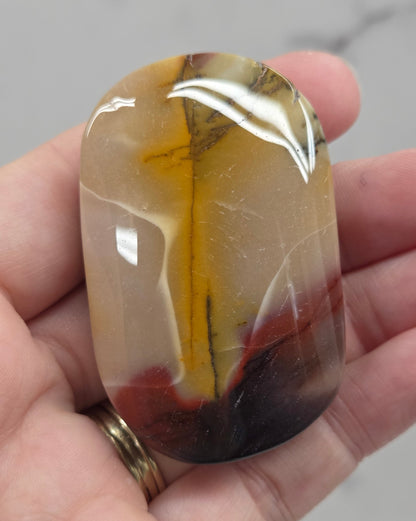A hand holding a polished, rounded agate stone with natural banding in shades of brown, yellow, and dark reddish-brown, set against a light gray marble background.