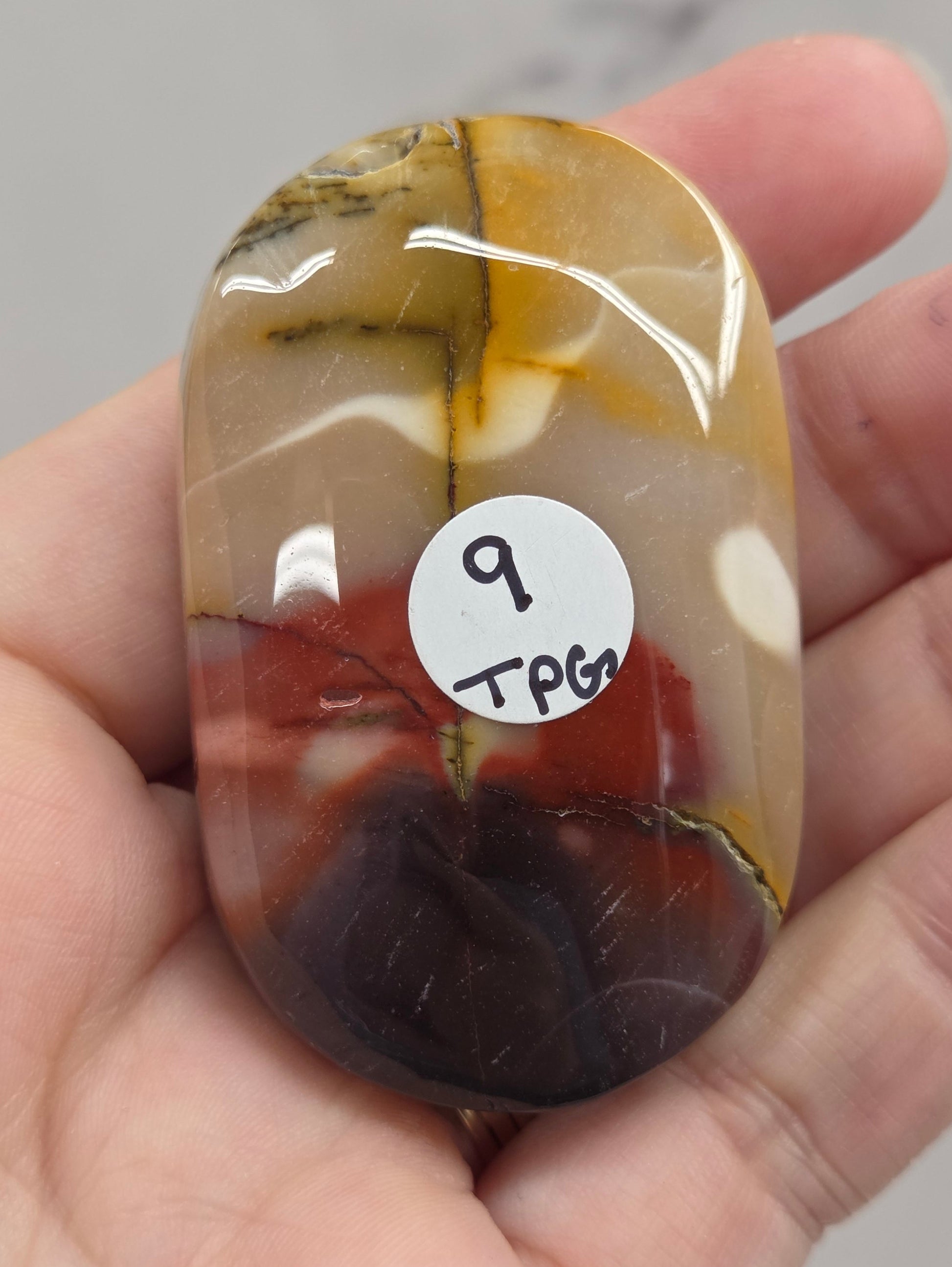 A hand holding a polished oval stone with marbled brown, amber, cream, and dark reddish-black patterns. A small white sticker with handwritten text is attached to the stone.