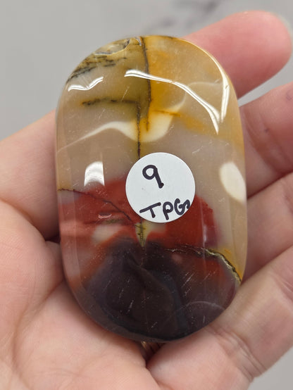 A hand holding a polished oval stone with marbled brown, amber, cream, and dark reddish-black patterns. A small white sticker with handwritten text is attached to the stone.