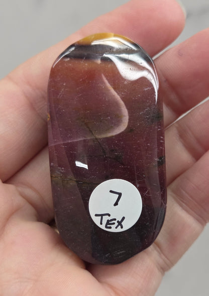 A polished oval gemstone with a marbled mix of dark purple, reddish-brown, and gold tones, held in a person's hand. A small white sticker with the text '7 Tex' is attached to the stone.