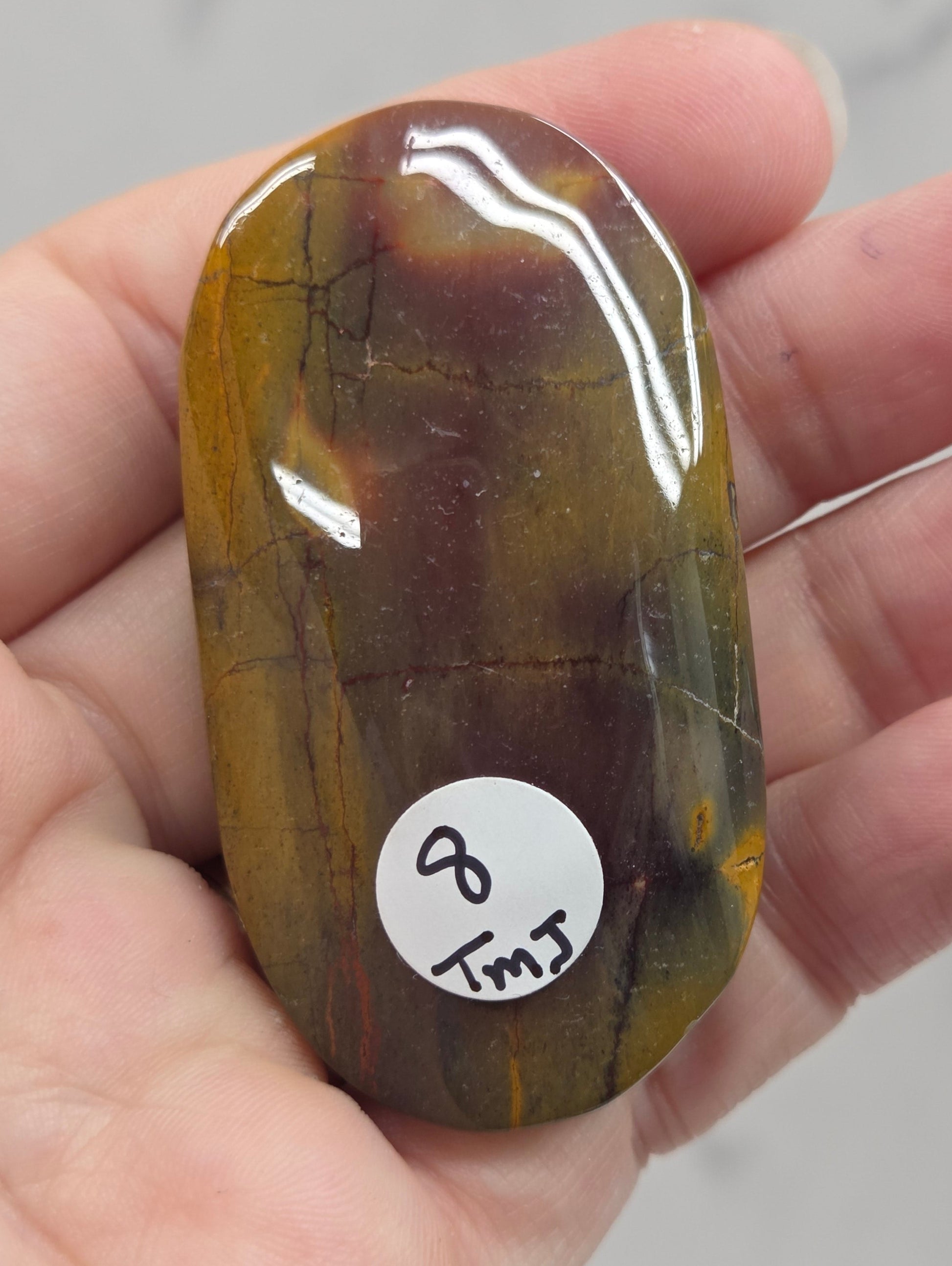 A hand holding a polished, smooth stone with a glossy finish, showing natural brown and yellow marbling. A small white circular sticker with handwritten text is attached to the stone.