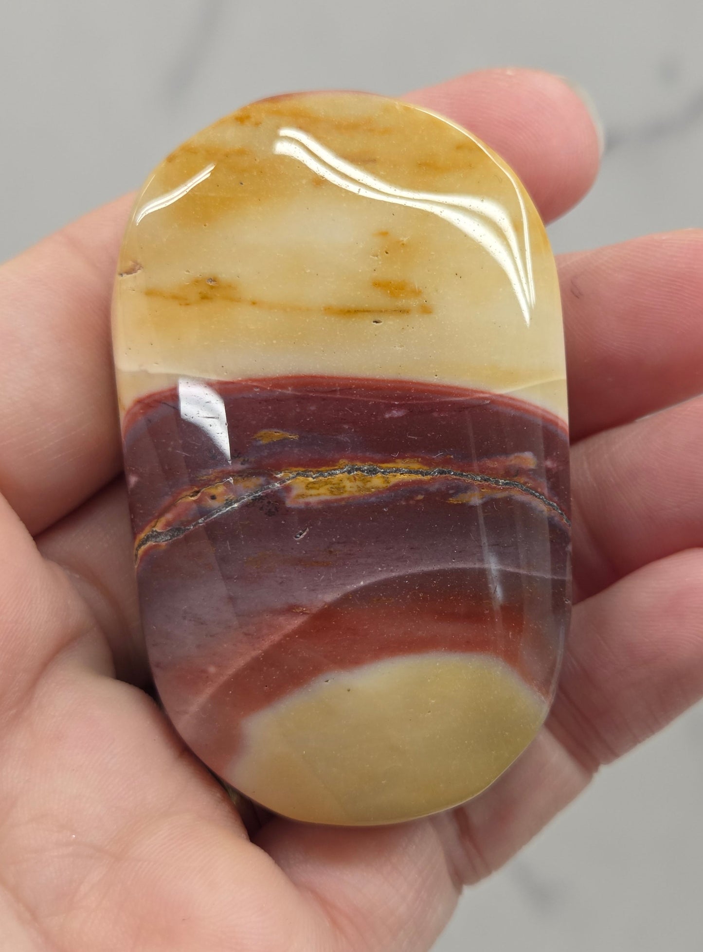 A polished stone with marbled cream, brown, and reddish-brown bands, held in a person's hand against a light gray marble background.