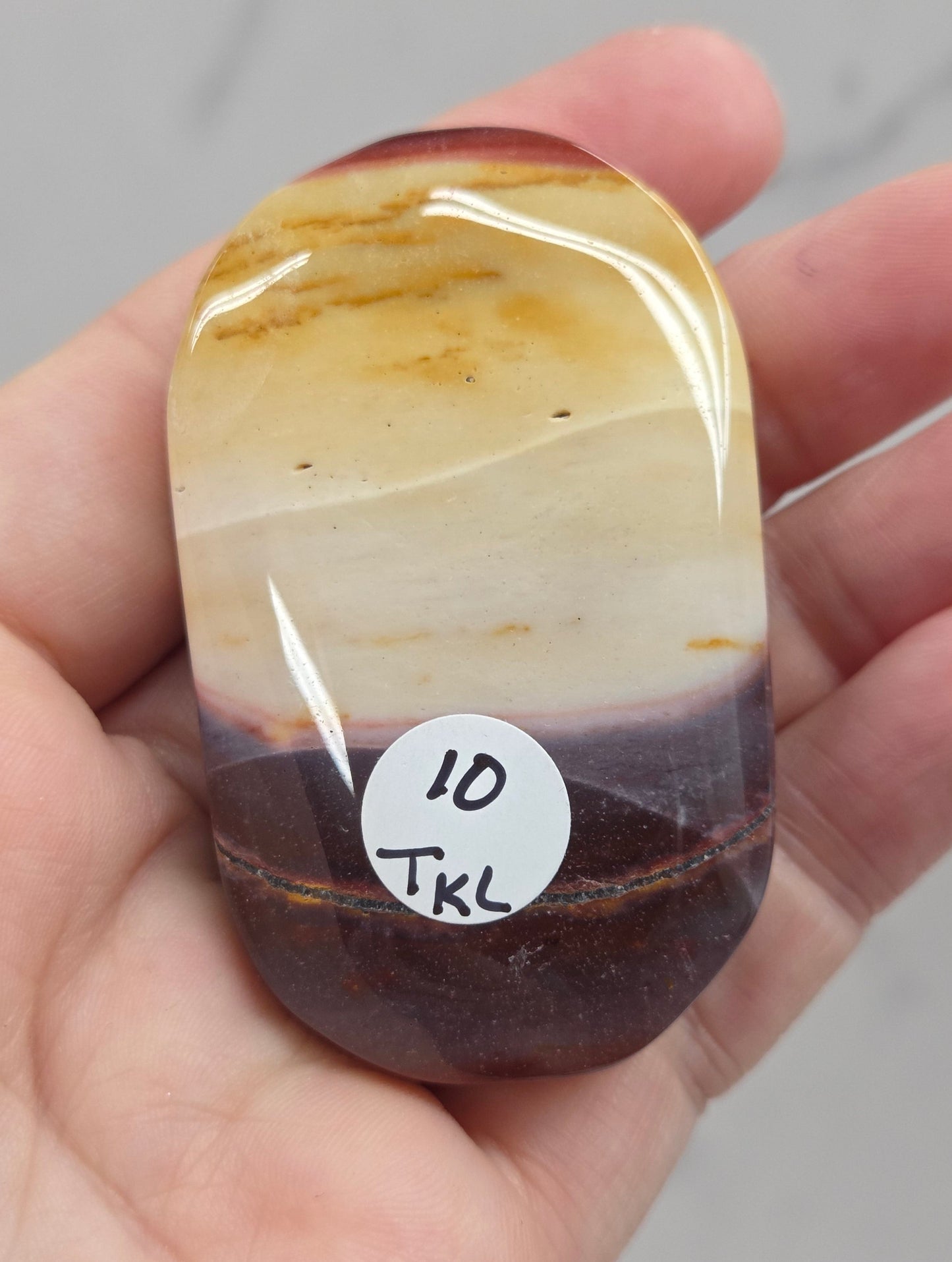 A hand holding a polished tumbled stone with a smooth rounded shape, featuring natural banding in cream, tan, and dark brown, with a small white sticker reading '10 TKL' on the side.