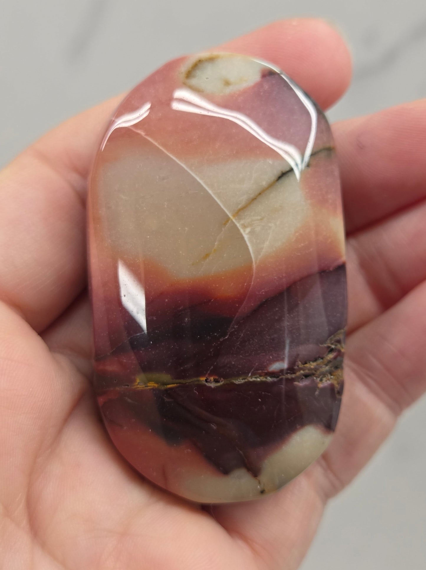 A polished stone with marbled brown, cream, and deep purple tones, held in a person's hand against a light gray background.