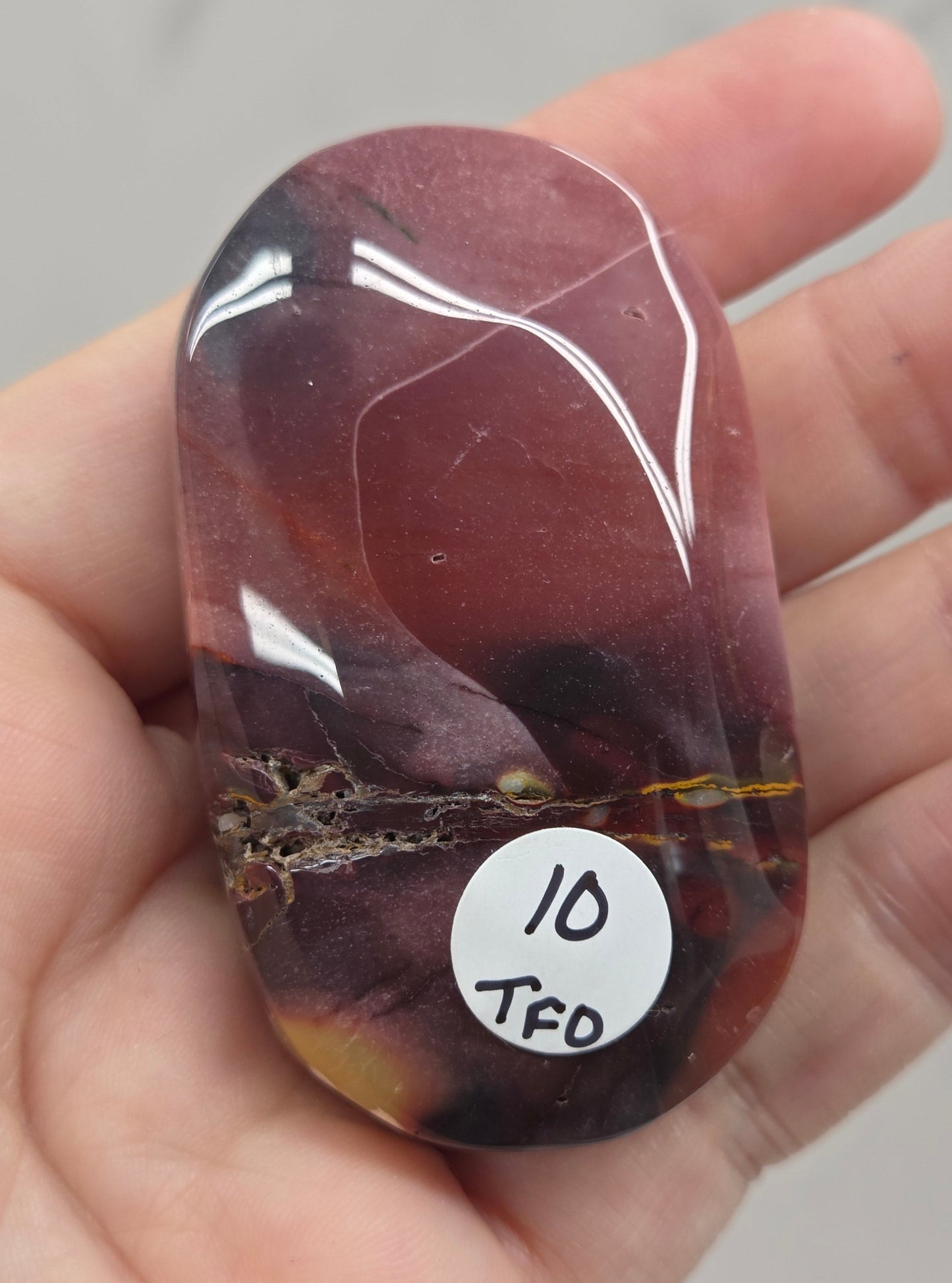 A hand holding a polished reddish-brown tumbled stone with a small white sticker that reads 'ID TFD'.