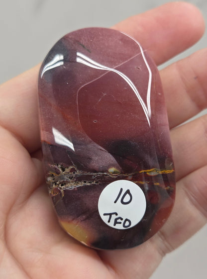 A hand holding a polished reddish-brown tumbled stone with a small white sticker that reads 'ID TFD'.