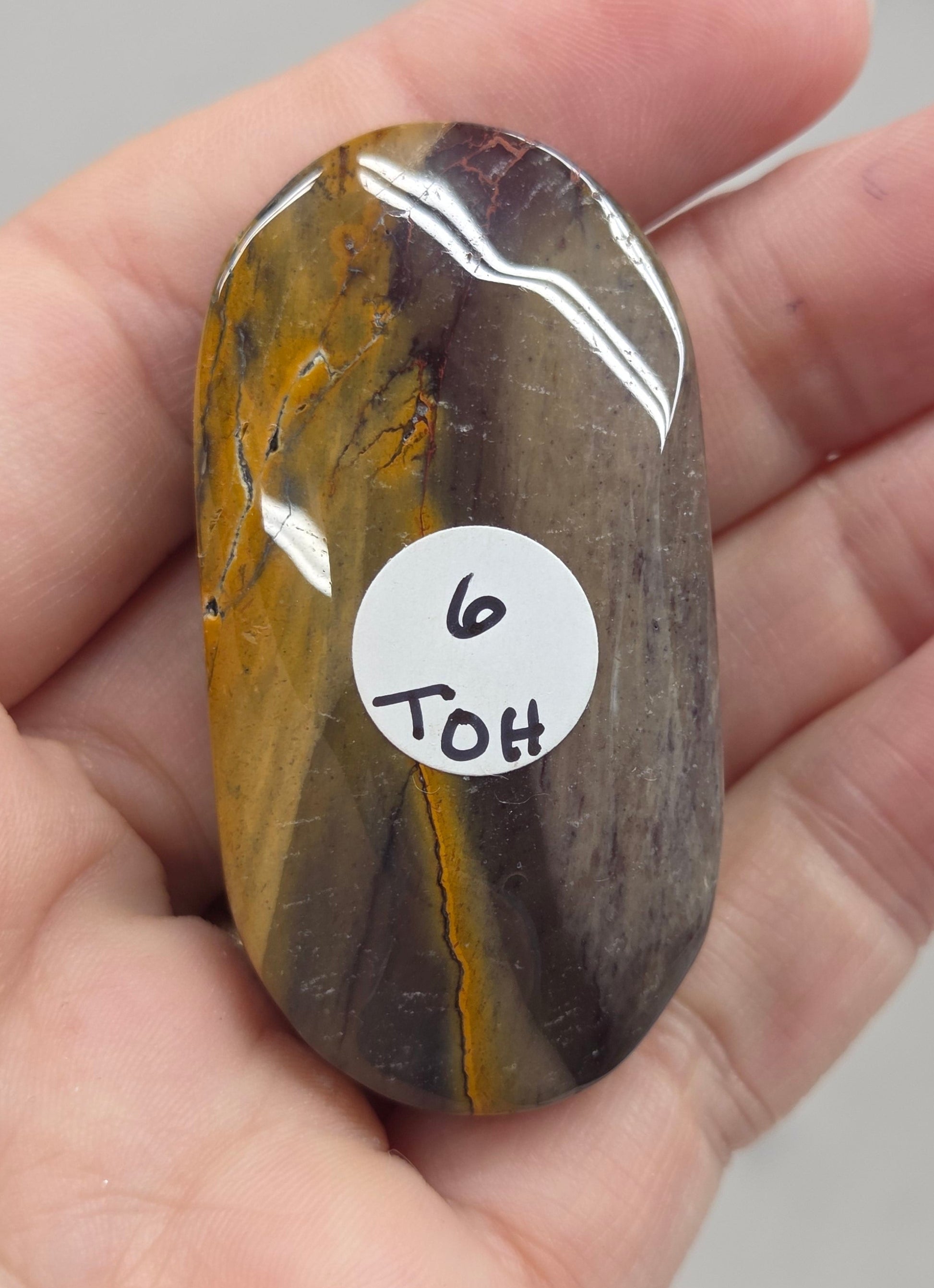 A polished mookaite stone held in a person's hand, showing a white circular sticker with the handwritten text '9 TOH'.