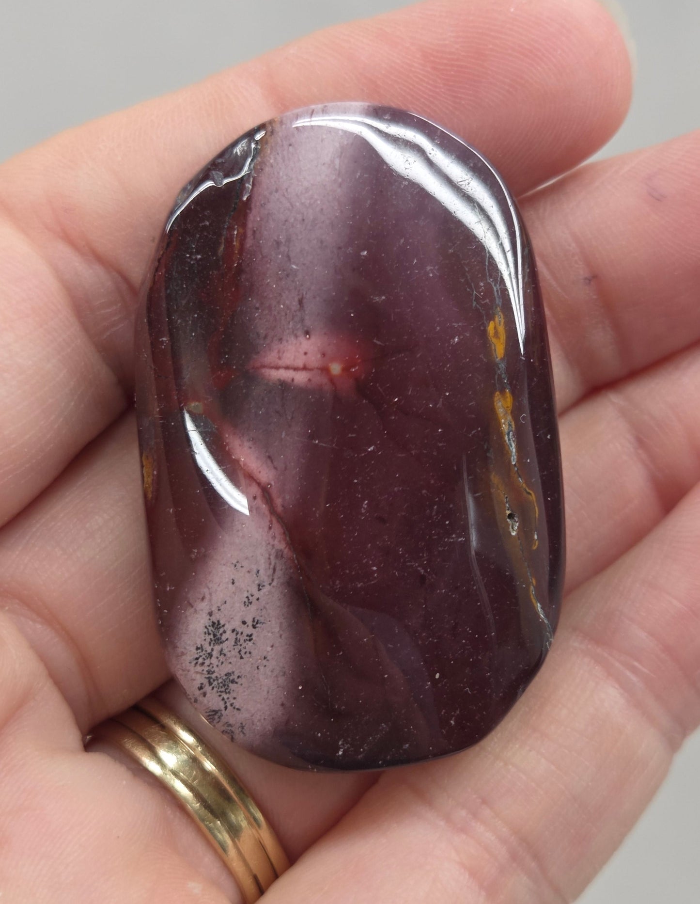 A polished tumbled gemstone with a dark purple and reddish-brown marbled pattern, held in a person's hand.
