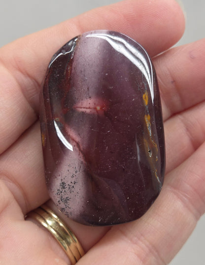A polished tumbled gemstone with a dark purple and reddish-brown marbled pattern, held in a person's hand.
