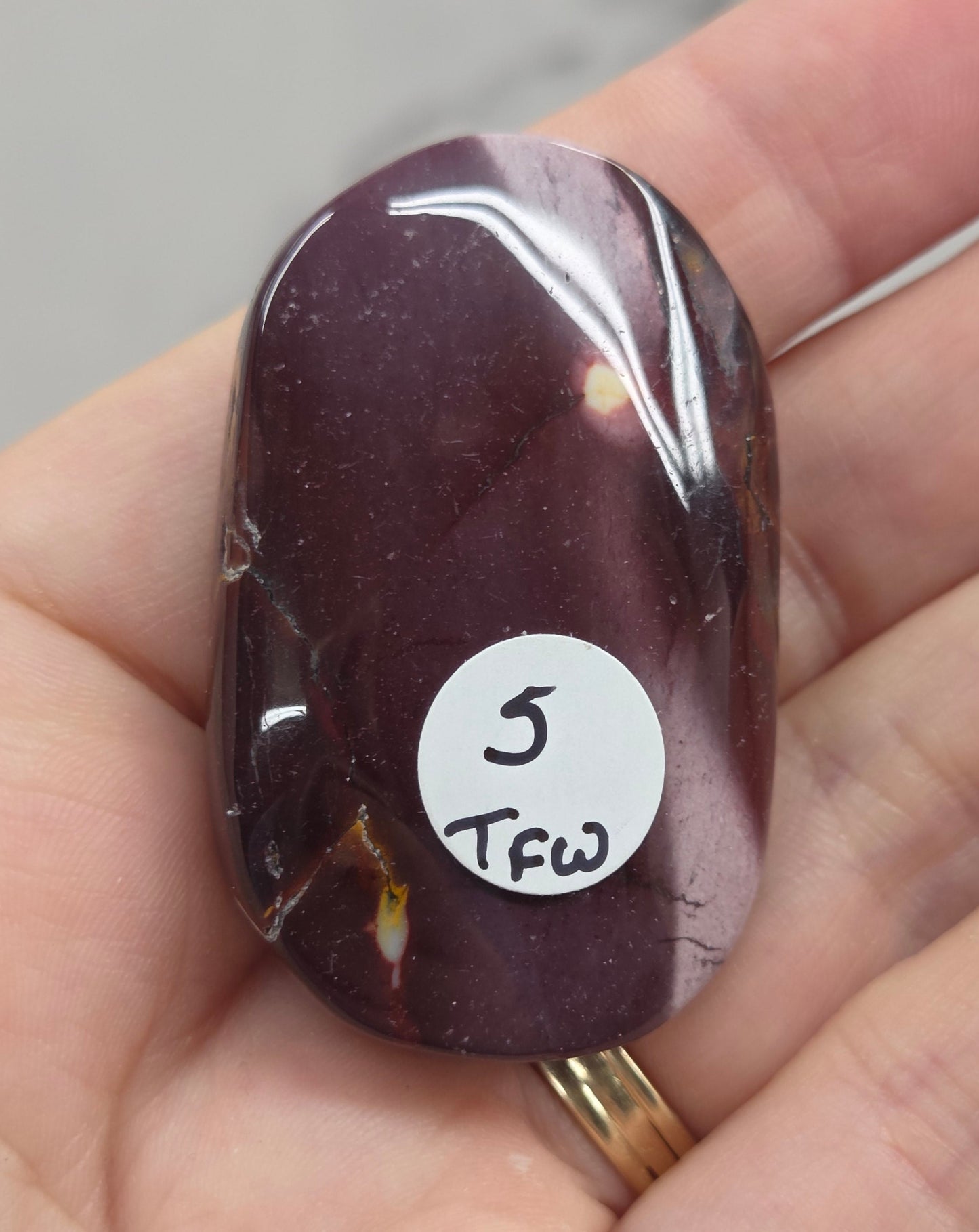 A polished dark purple tumbled stone pendant or charm held in a person's hand. The stone has a small white circular sticker on it with handwritten text '5 TFW'.