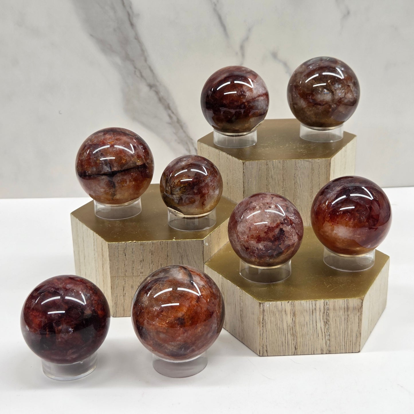 Eight polished brown and reddish stone spheres displayed on small clear stands, arranged on light wood hexagonal pedestals against a white marble-look background.