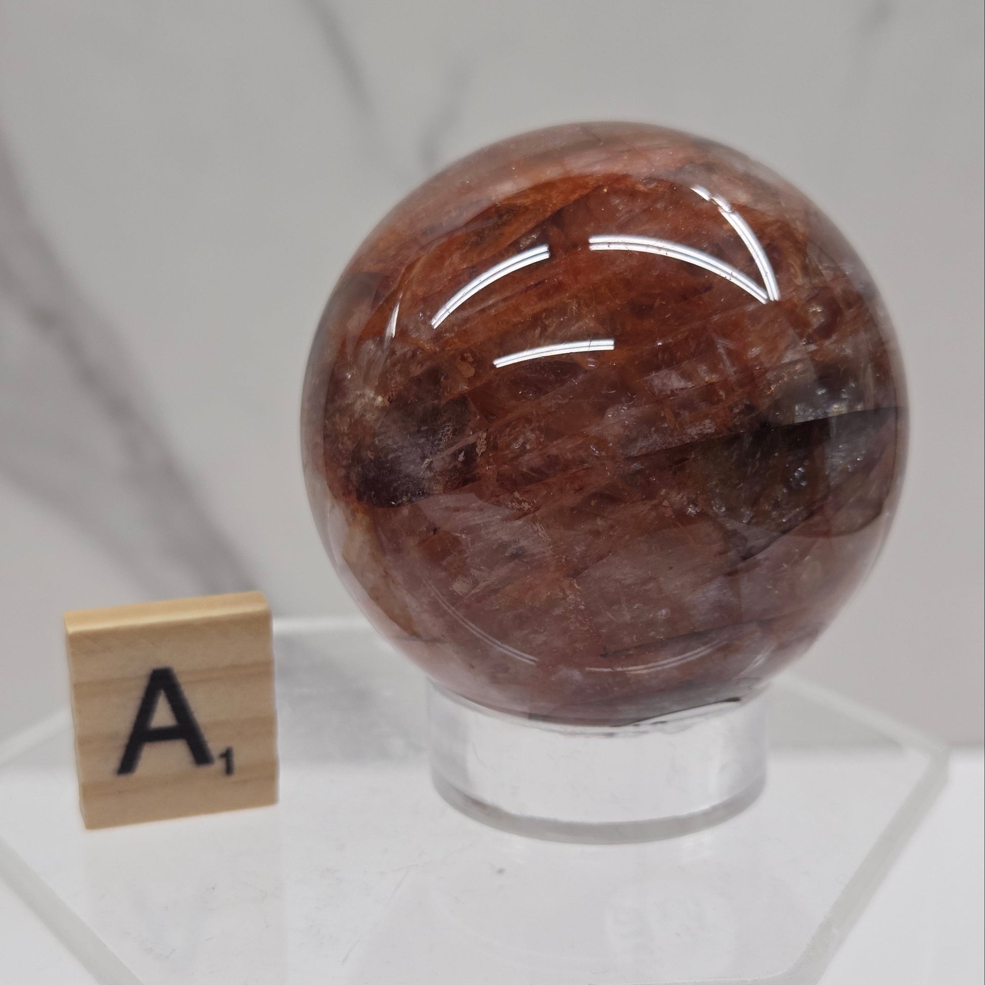 A polished brown gemstone sphere mounted on a clear acrylic stand, with a small letter tile marked 'A1' below it, set against a white marble background.
