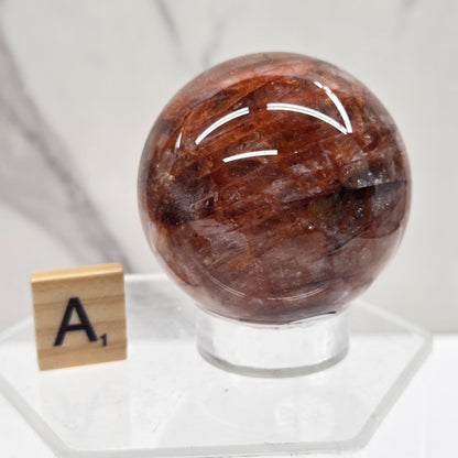 A polished brown gemstone sphere displayed on a clear acrylic stand, with a small wooden letter tile showing the letter 'A' nearby, set against a white marble background.