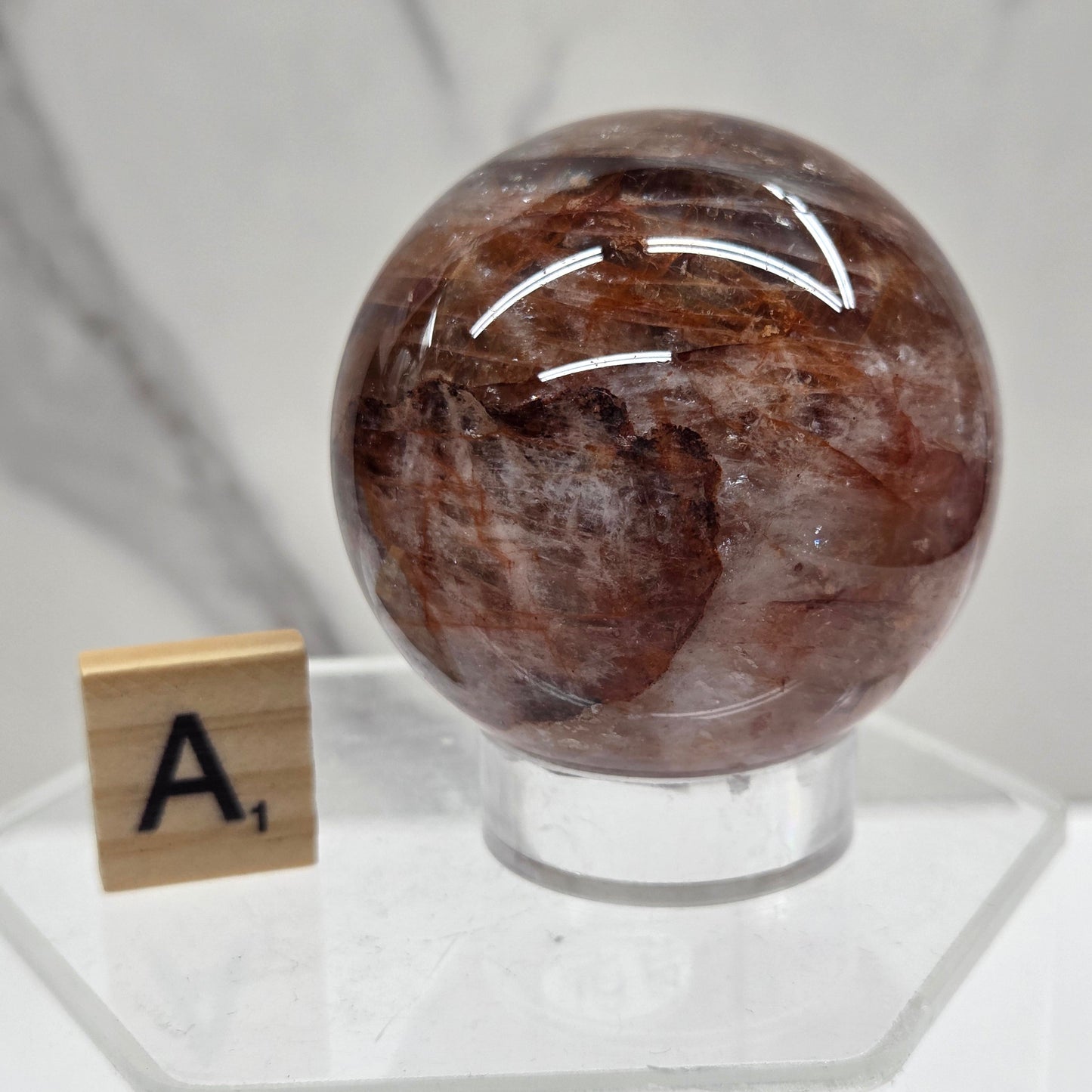 A polished brown stone sphere mounted on a clear acrylic stand, photographed against a white marble-like surface with a small letter tile near the base.