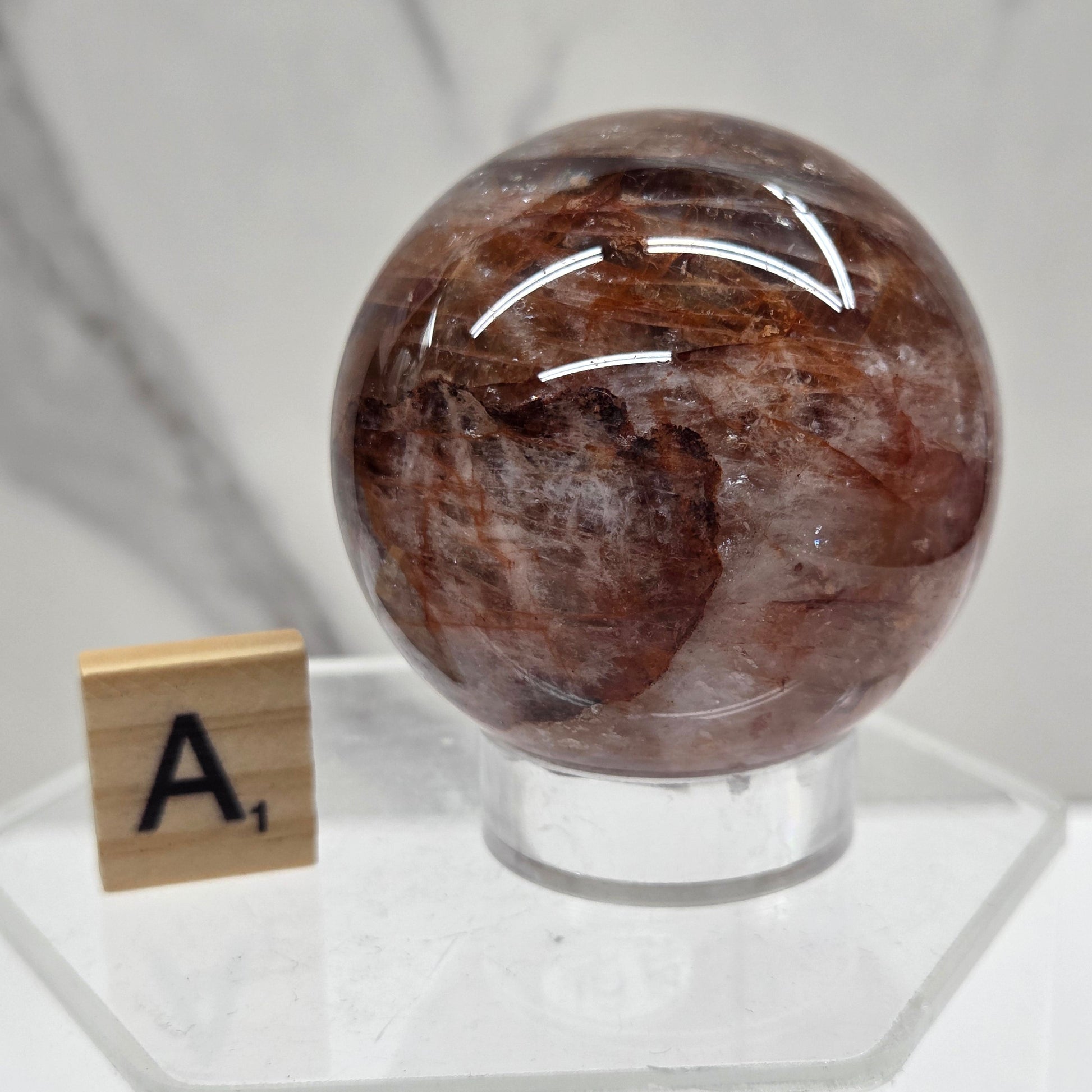 A polished brown stone sphere mounted on a clear acrylic stand, photographed against a white marble-like surface with a small letter tile near the base.