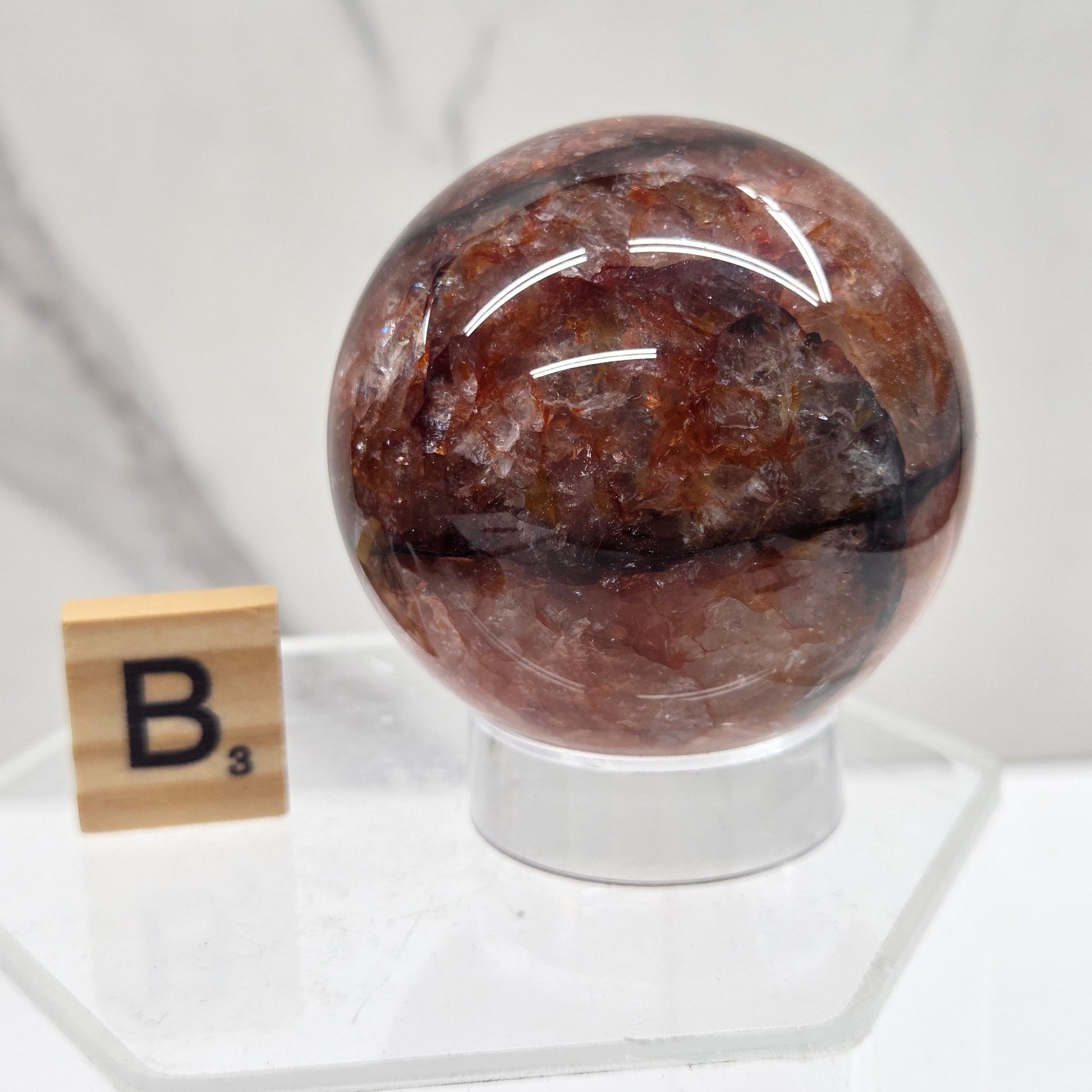 A polished brown gemstone orb with natural inclusions, mounted on a clear acrylic stand next to a small wooden tile labeled B3, set against a white marble background.