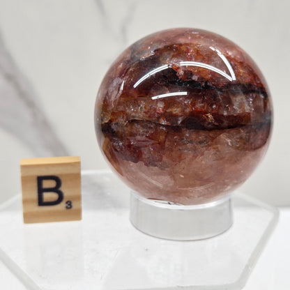 A polished brown crystal sphere with dark inclusions, mounted on a clear acrylic stand with a small wooden tile labeled 'B' nearby, set against a white marble background.
