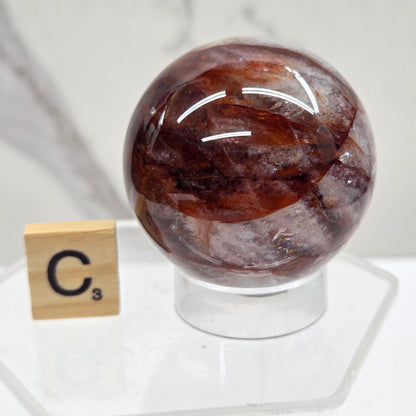 A polished brown and smoky gray stone sphere mounted on a clear acrylic stand, with a small wooden tile showing 'C³' placed below it, against a light marble-like background.