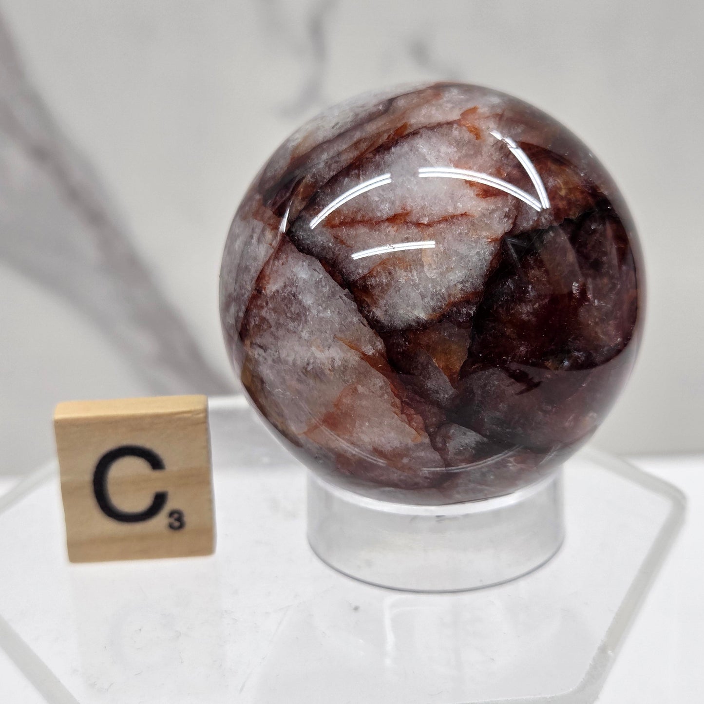 A polished brown and white marbled gemstone sphere displayed on a clear acrylic stand next to a small wooden tile labeled C3, set against a white marble background.