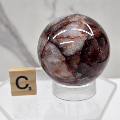 A polished brown and white marbled gemstone sphere displayed on a clear acrylic stand next to a small wooden tile labeled C3, set against a white marble background.