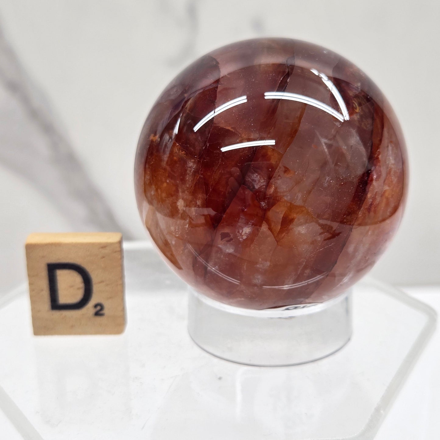 A polished brown gemstone sphere displayed on a clear acrylic stand, with a small wooden letter tile showing the letter 'D' nearby, set against a white marble background.