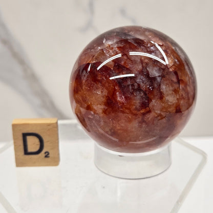 A polished brown gemstone sphere displayed on a clear acrylic stand with a small wooden tile labeled 'Dm' nearby, set against a white marble-patterned background.