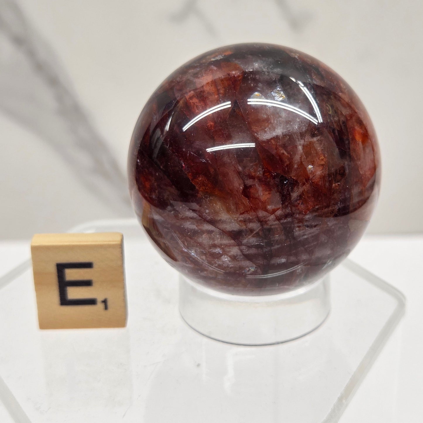 A polished round gemstone with a mottled dark brown and reddish-brown pattern, mounted on a clear acrylic stand with a small gold tile nearby for scale, set against a white marble-patterned background.