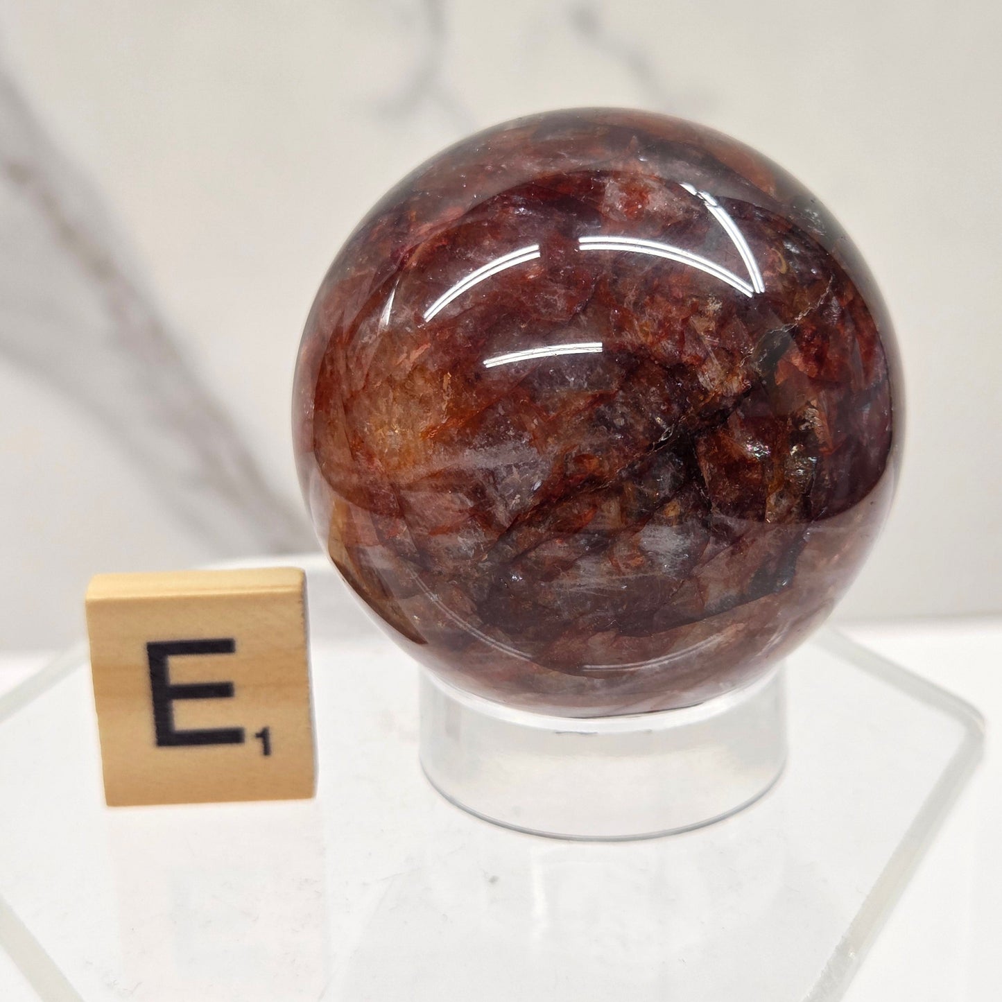 A polished round brown gemstone or crystal sphere mounted on a clear acrylic stand, with a small wooden block displaying the Hebrew letter Shin in front, set against a white marble-patterned background.