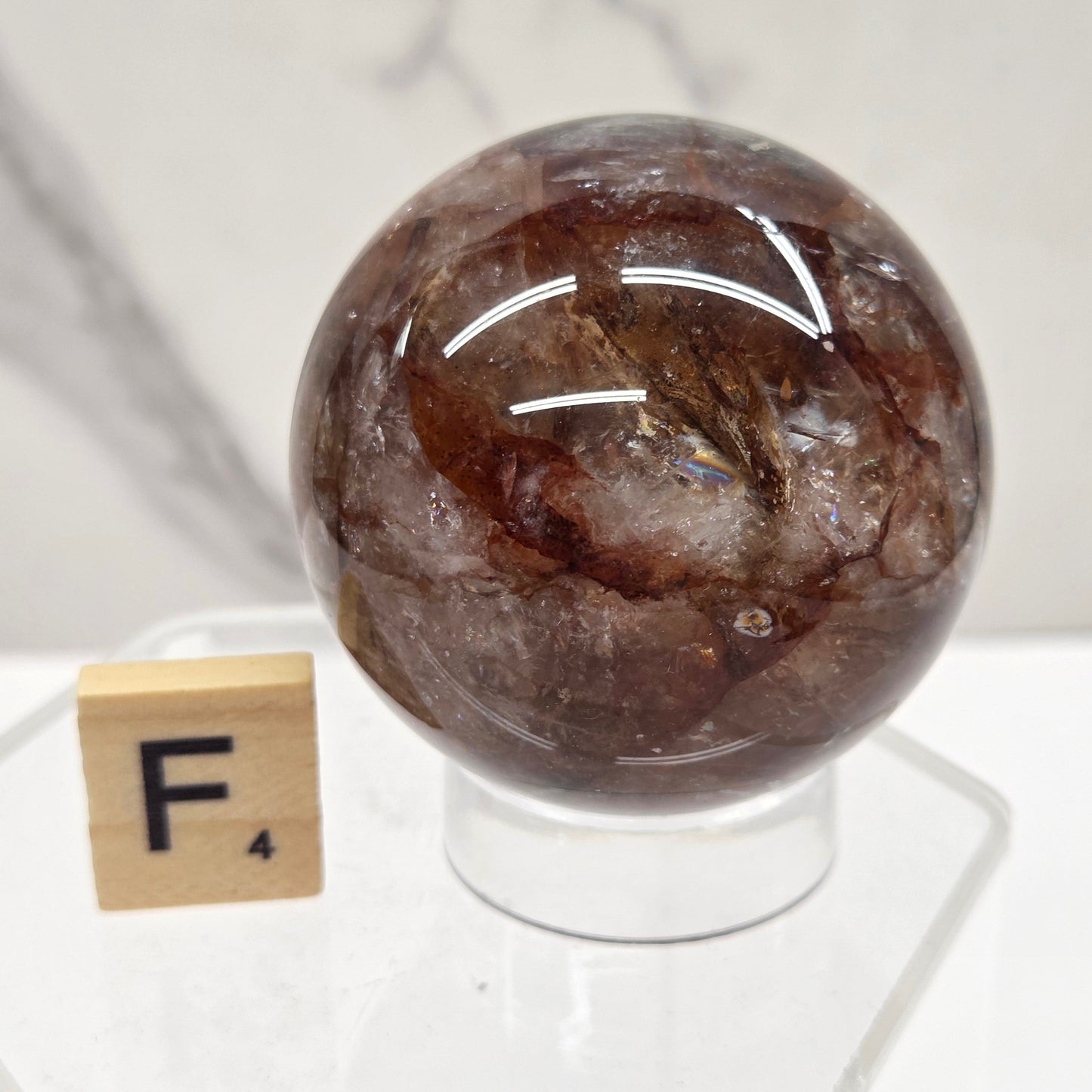 A polished brown gemstone sphere mounted on a clear acrylic stand, photographed against a white marble-like surface with a small wooden tile nearby.