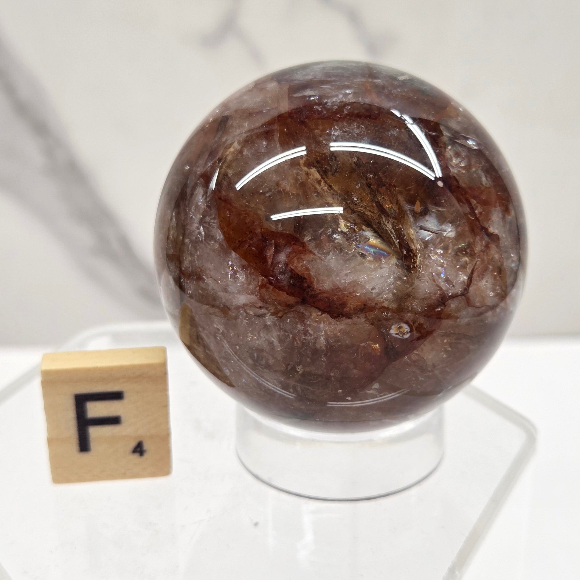 A polished brown gemstone sphere mounted on a clear acrylic stand, photographed against a white marble-like surface with a small wooden tile nearby.