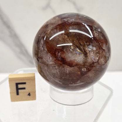 A polished brown gemstone sphere mounted on a clear acrylic stand, photographed against a white marble-like surface with a small wooden tile nearby.