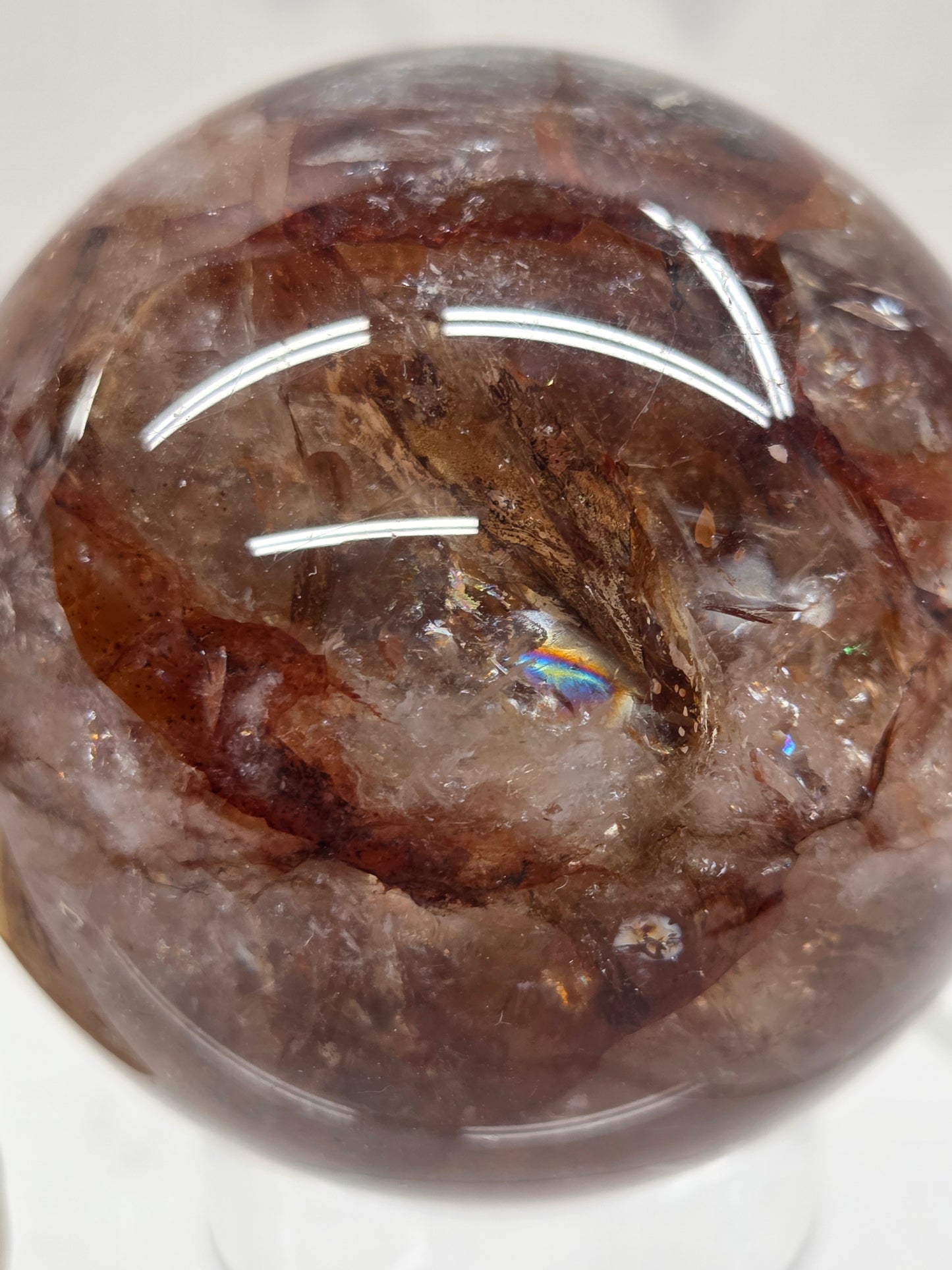 A close-up of a polished brown gemstone sphere with natural inclusions and a small iridescent rainbow flash reflection in the center, photographed on a white background.