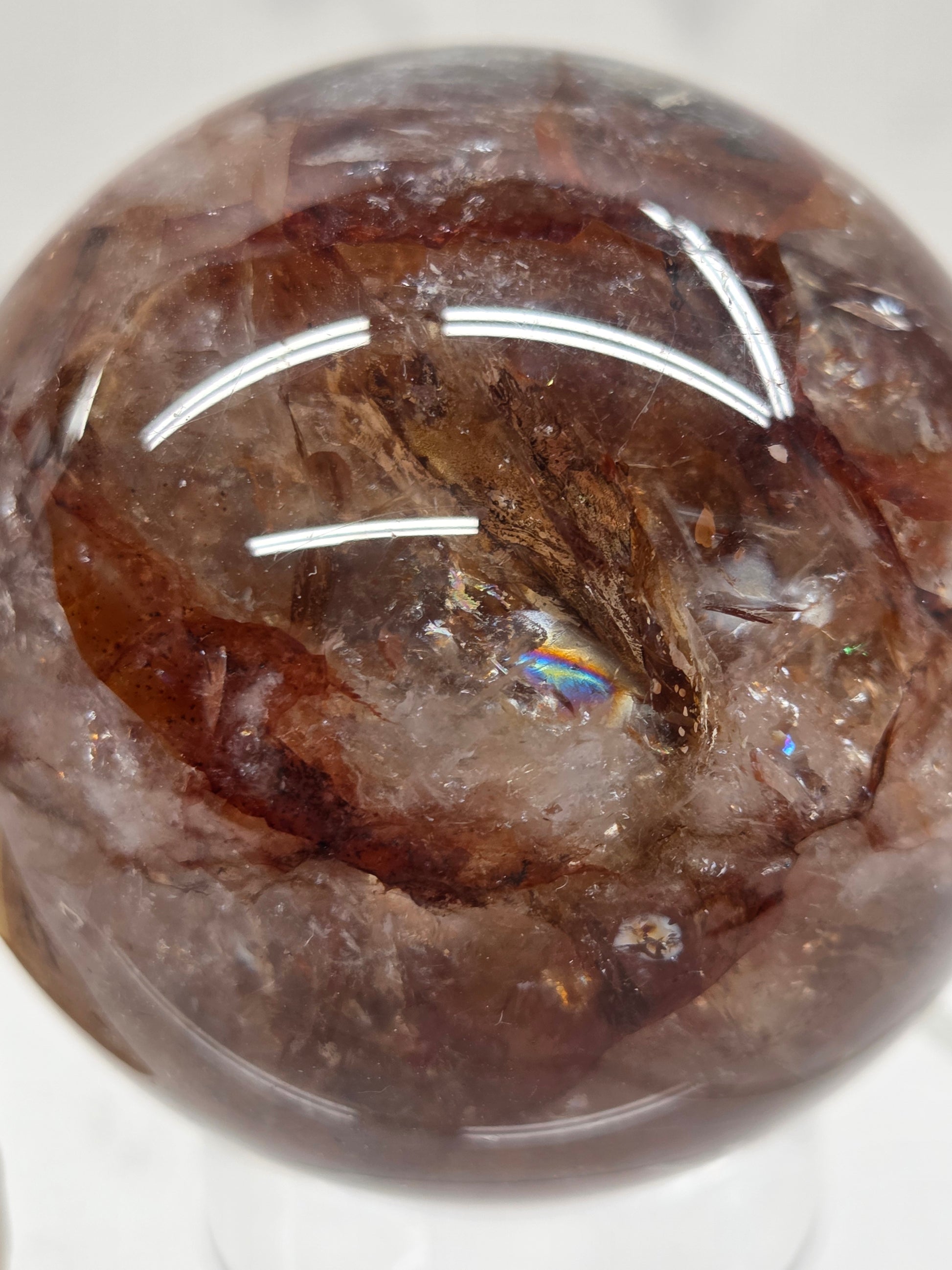 A close-up of a polished brown gemstone sphere with natural inclusions and a small iridescent rainbow flash reflection in the center, photographed on a white background.