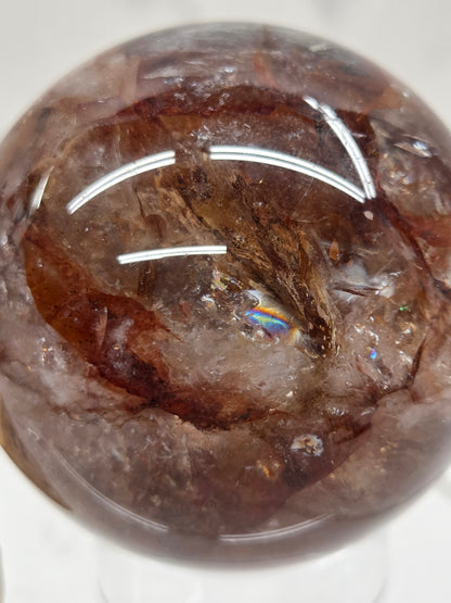A close-up of a polished brown gemstone sphere with natural inclusions and a small iridescent rainbow flash reflection in the center, photographed on a white background.