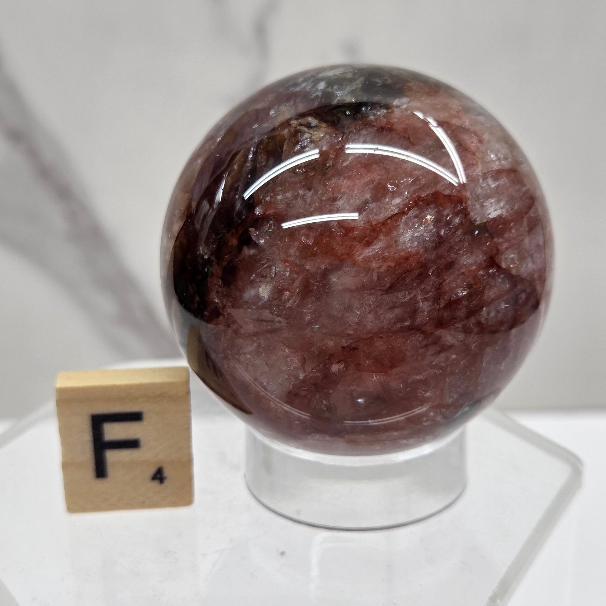 A polished round brown stone sphere mounted on a clear acrylic stand with a small wooden letter tile below it, photographed against a light marble-like surface.