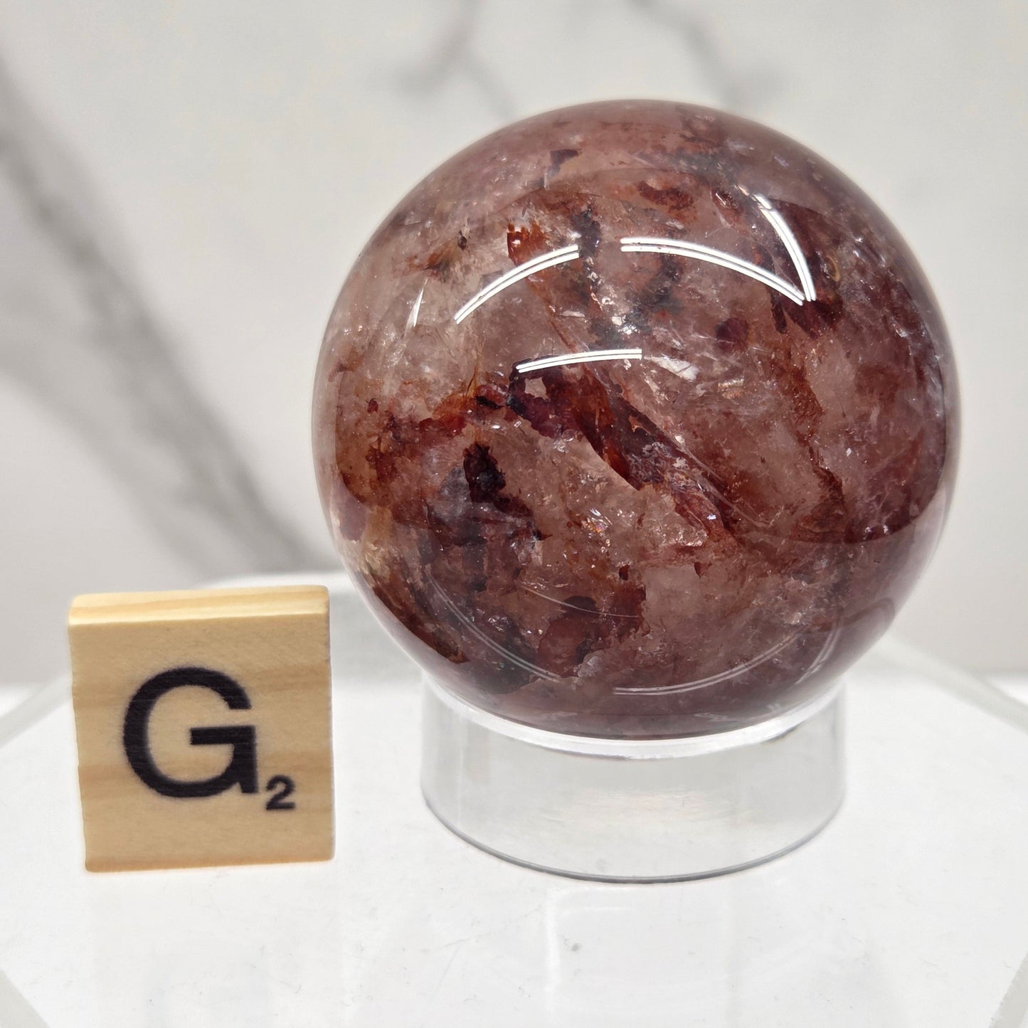 A polished brown crystal sphere displayed on a clear acrylic stand, with a small Scrabble tile showing the letter G placed below it, against a white marble background.